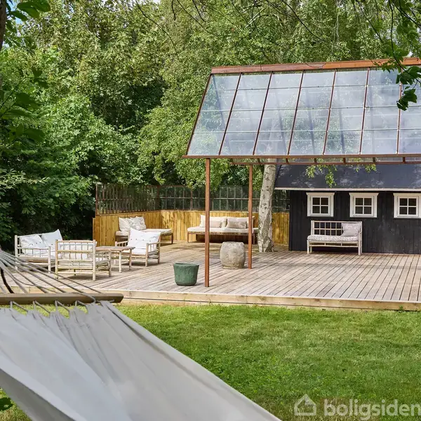 Hængekøje hænger mellem træer, ved siden af terrasse med sofagruppe og gennemsigtigt tag. I baggrunden står hus omgivet af frodige træer.