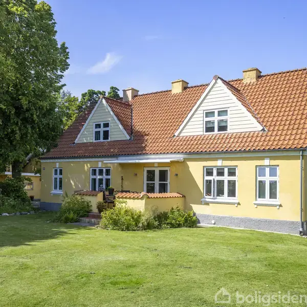 Et gult hus med rødt tegltag står i en velholdt have. Flere vinduer og en indgangsdør er synlige. Til venstre er der store træer, og en blå himmel ses ovenover.
