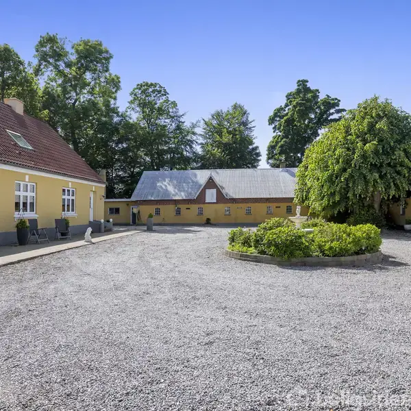 Gule bygninger står omkring en gårdsplads med grus. Der er en grøn busk i midten, og træer omgiver området under en klar blå himmel.