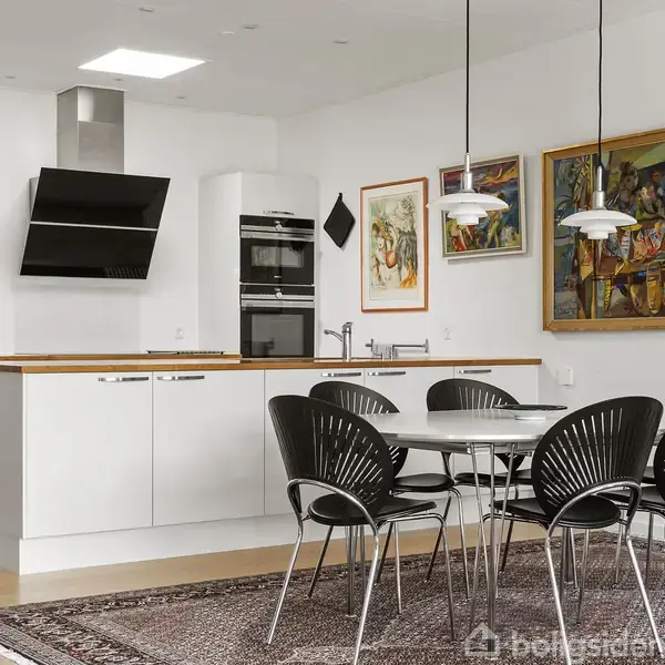 Køkken med hvidt køkkenbord og træbordplade, sort emhætte, stålkomfur, og sort spisebord med seks stole. Omgivet af indrammede malerier på hvide vægge og trægulv.