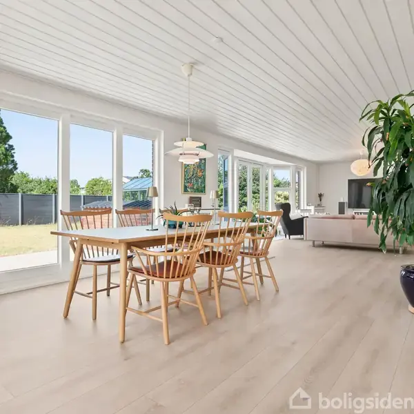 Spisebord med træstole står i en lys stue med glasdør mod haven. En stor grøn plante i hjørne, og sofa i baggrunden. Elegant belysning hænger over bordet.
