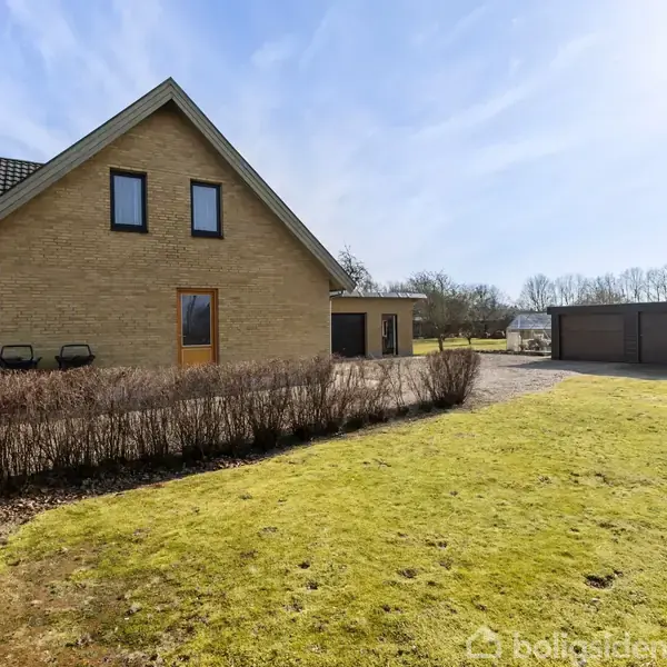 Hus med gult murstensfacade står stille i en grøn have med hække. I baggrunden findes en garage ved grusvej. Himlen er klar og solrig.