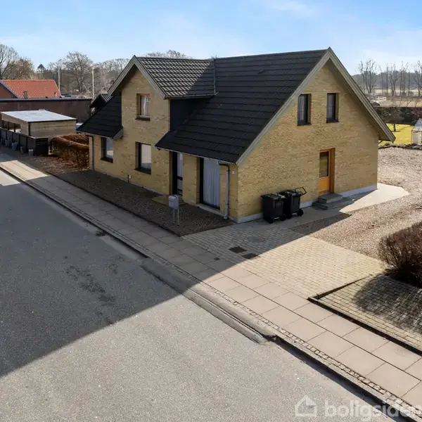 Et gulstenshus med sort tag står ved en stille villavej. Der er bede og fortov omkring huset, og området er omgivet af grønne marker og enkelte træer i baggrunden.