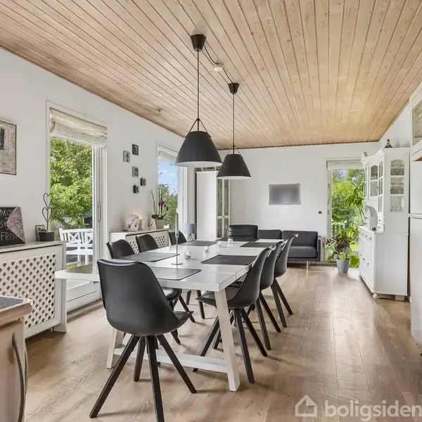 Et spisebord med sorte stole står i en lys, moderne spisestue. Rummet har træloft, store vinduer og udsigt til en have. Der hænger tre sorte lamper over bordet.