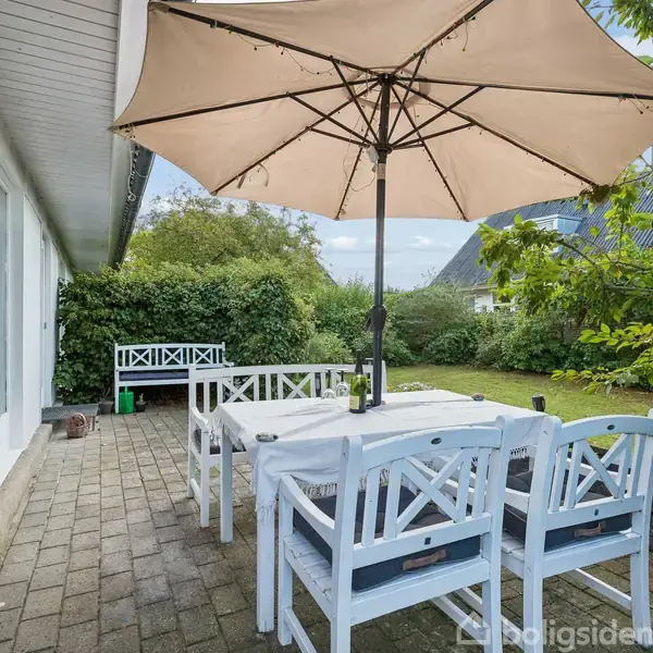 En hvid havestue står under en stor parasol på en terrasse med flisebelægning, omgivet af grønne buske og en velplejet græsplæne i en have.