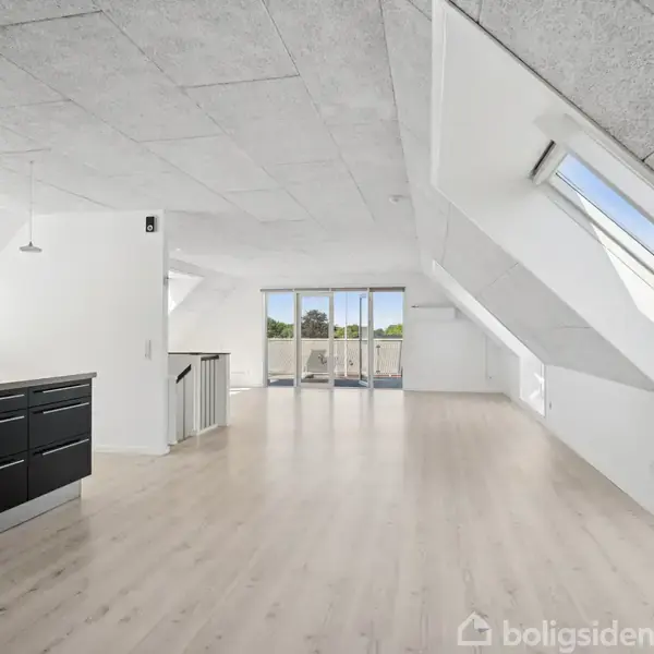 Et lyst, rummeligt loftsrum med skråvinduer og et elegant sort køkkenmodul står tomt. Store glasdøre fører til en altan, og lyse trægulve dækker hele rummet.