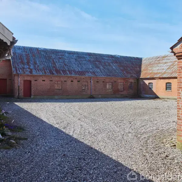 Bygning af røde mursten med tag i bølgeblik står stille i en gårdsplads dækket af grus. Sollys skaber skygger på jorden. Skilt med tekst "LILLEGÅRD" ses på væggen.
