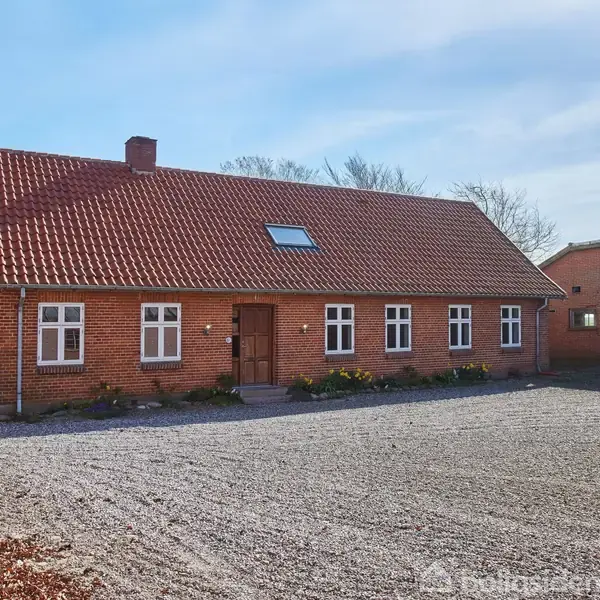 En rødstensbygning med rødt tegltag står stille, omgivet af en grusbelagt gårdsplads. Der er flere vinduer på facaden og en enkelt dør i midten.