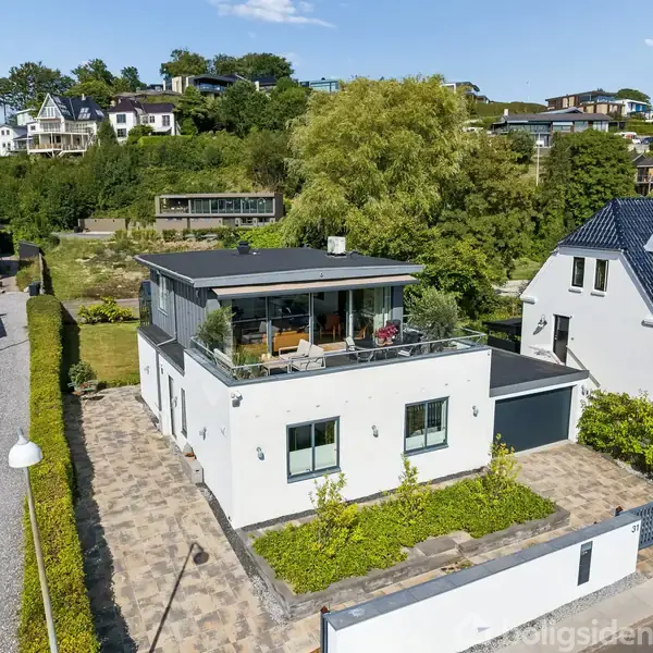 Et moderne, hvidt hus med en stor terrasse på første sal, omkranset af grønne hække og træer, står på en flisebelagt grund i et boligområde med andre huse i baggrunden.