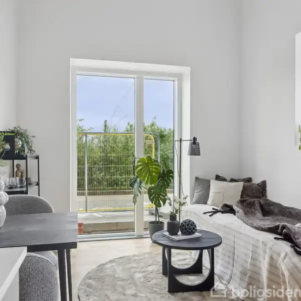 Stue med sofabord og sofa, dækket af et tæppe. Et rundt tæppe ligger på gulvet. Store vinduer åbner ud mod en balkon med udsigt til grønne træer. Plakater og planter pynter væggene.