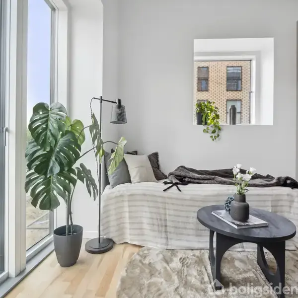 En enkeltseng dækket med en stribet tæppe står ved siden af et stort vindue. Ved siden af, en gulvlampe og en plante. Et spejl hænger på væggen.