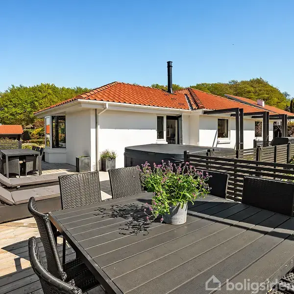 Hus med rødt tegltag har en stor terrasse med havemøbler og planter. Omkranset af grønne træer og andre huse, i et solrigt og fredeligt boligområde.