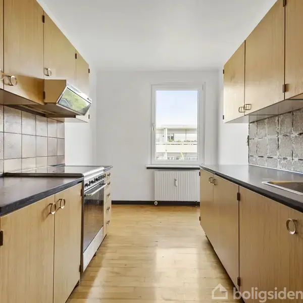 Køkken med træskabe og sort bordplade, moderne komfur centralt placeret, vindue giver naturligt lys, væg med brune fliser. Radiator under vinduet. Ingen tekst fundet.