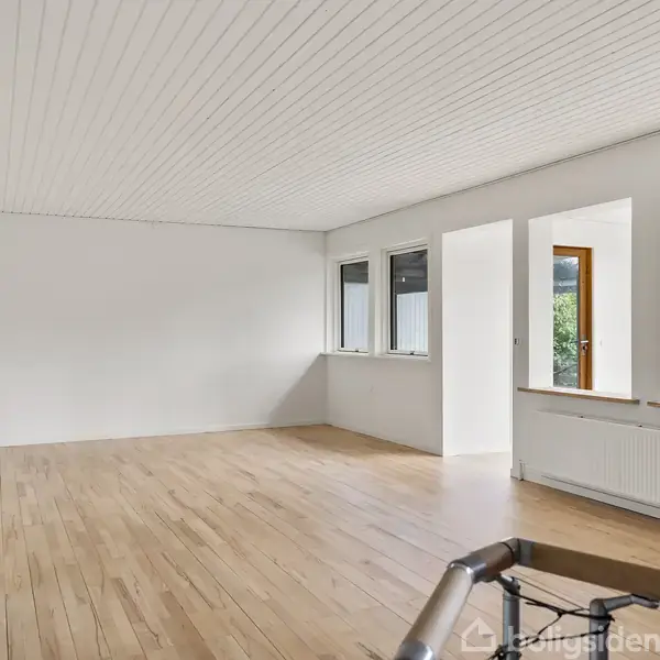 Et lyst, tomt rum med trægulv og hvidt loft. Flere vinduer og en dør åbner mod grønne omgivelser. En radiator er placeret under vinduerne.