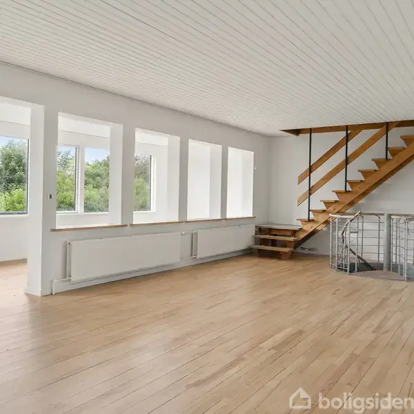 Et åbent rum med trægulv indeholder en trappe, der leder opad. Store vinduer fylder væggen og giver udsigt til en grøn have. Væggene og loftet er hvide.