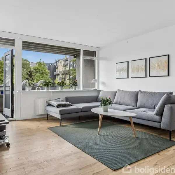Sofa står i en stue nær et stort vindue med udsigt til grønne træer og bygninger. Ved siden af sofaen er et sofabord og malerier hænger på væggen.