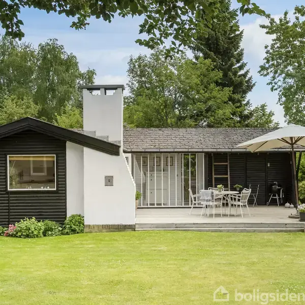 Boliger omgivet af frodige træer ligger i et grønt kvarter nær havet. På nogle huse ses røde tage og velplejede haver. Baggrunden viser kysten og havet i horisonten.