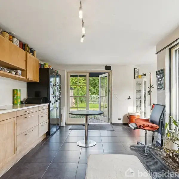 Køkken med træskabslåger, vask og et bord i midten, åbne døre fører til grøn have, og vinduer lader naturligt lys strømme ind. Planter placeret ved vindueskarmen.