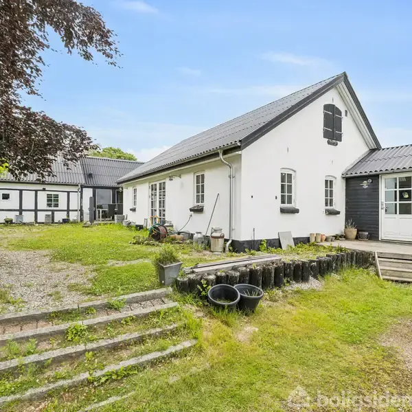 Huset er hvidt med sort bindingsværk, omgivet af en have og en grussti. Trætrappe fører til en græsplæne. Blå himmel over scenen.