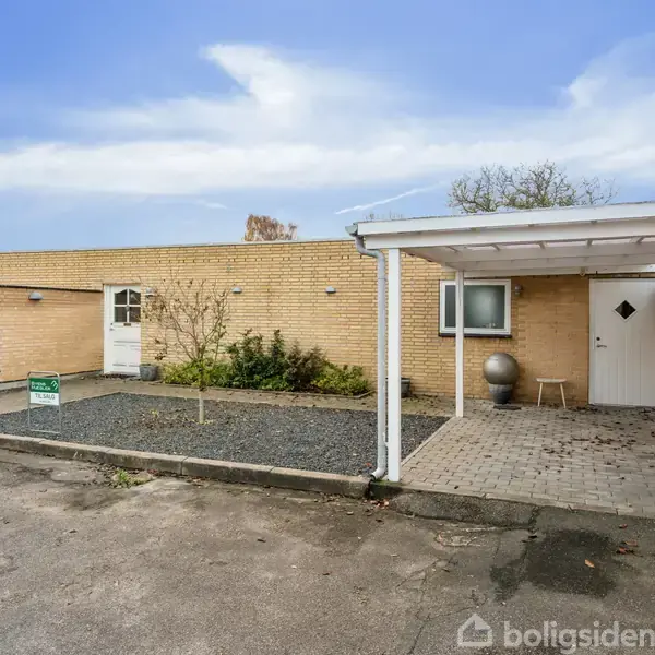 Et fladt, gulstenshus med en carport ved indgangen. Omkringliggende fliser er delvis dækket af småsten og planter. Skyfri blå himmel. En postkasse og et skilt er placeret til venstre.