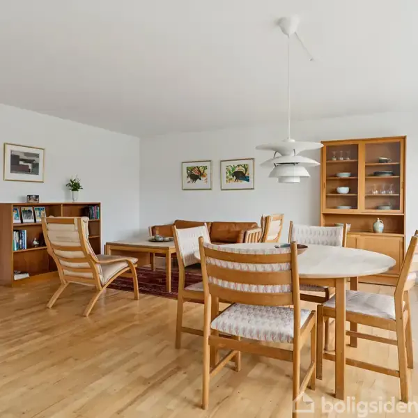 Et spisebord med seks stole står i et lyst stueområde med trægulv. Omgivet af en reol med bøger, vægkunst, og et glasskab. Store vinduer til højre.