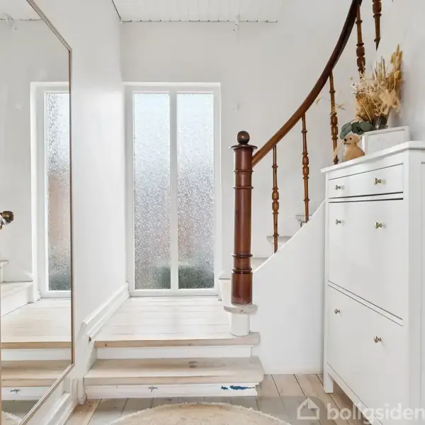En hvid trappe med trægelænder fører opad i en lys entre med stort spejl og skoskab. Omkring trappen står dekorationer på skabet, og en glasdør giver naturligt lys.