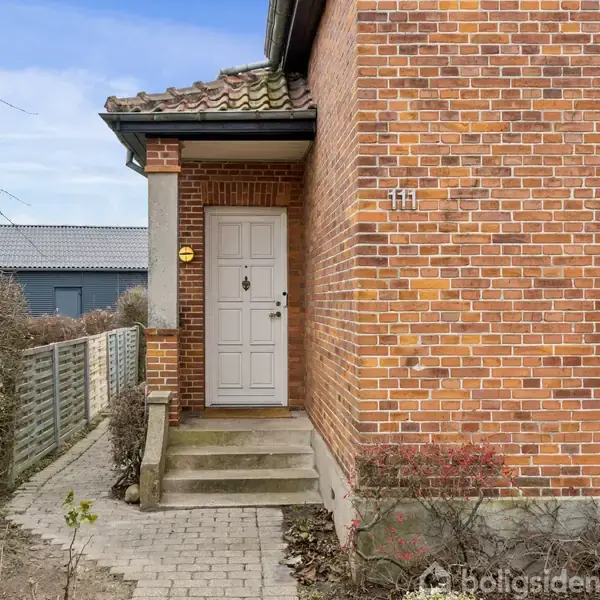 Murstenshus' indgang med hvid dør findes øverst på en trappe. Omgivet af en flisebelagt sti og have. Nummeret "111" ses på muren ved siden af døren.