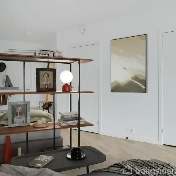 Bogreol med billeder, lampe og bøger står i et værelse med lyst trægulv. I baggrunden ses en seng og to lukkede, hvide døre samt et abstrakt maleri på væggen.