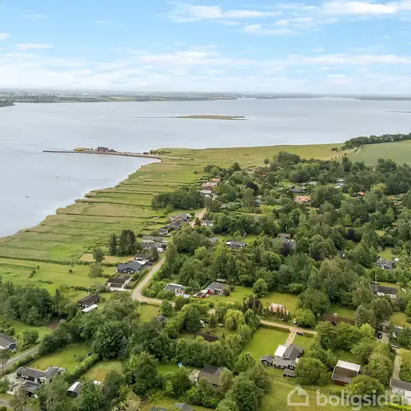 Kystnære huse ligger spredt i et frodigt, grønt område ved en stor sø eller fjord. Landskabet er præget af skov og marker, med en lang kystlinje i baggrunden.