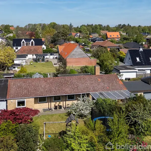 En etplans villa med rødt tegltag står i en velplejet have. Trampolin og gyngestativ i forgrunden. Omkringliggende parcelhuskvarter med grønne træer og farverige tage.