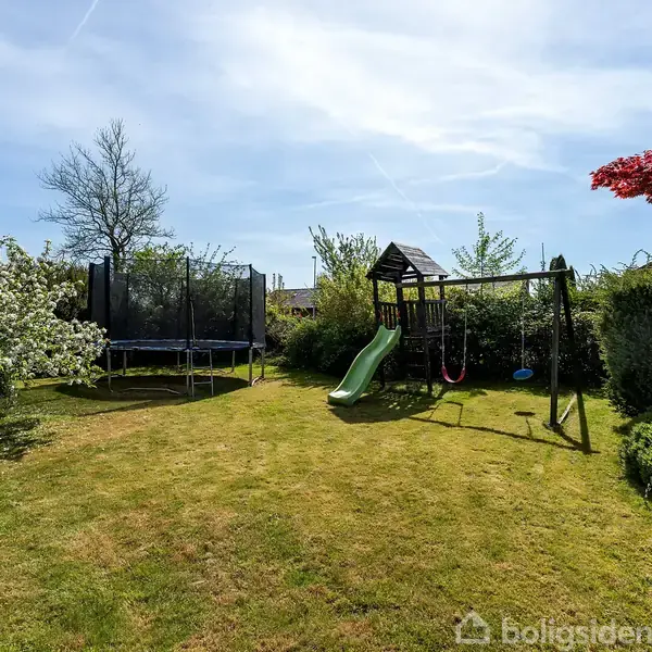 En trampolin står på en græsplæne i en have, omgivet af træer og buske. Til højre ses et legetårn med en rutsjebane og gynger.