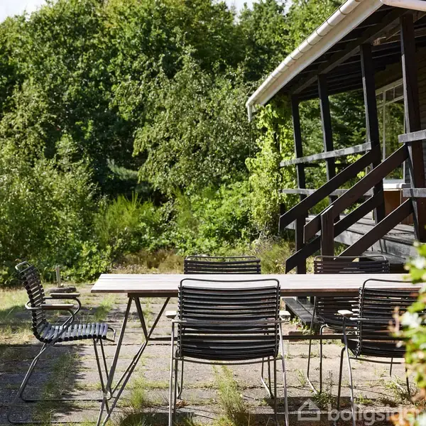En langbord med sorte metalstole står på en terrasse foran et træhus med en veranda, omgivet af tæt grøn bevoksning.