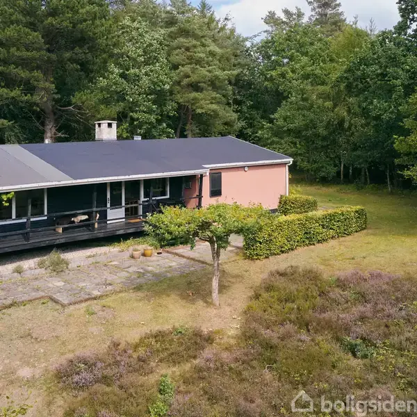 Et en-etagers hus med sort tag og terrasse omkranset af grønne træer. Foran ligger en gårdhave med stenbelægning. Vegetation og buske omgiver ejendommen.