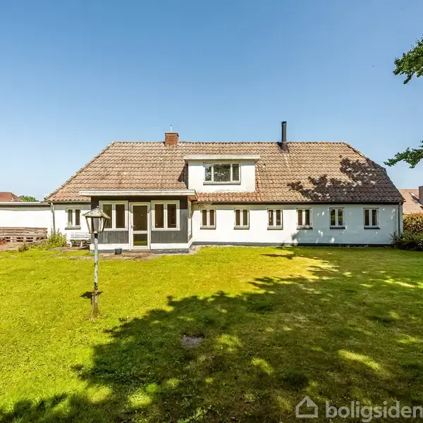 Et hvidt hus med brunligt tag står på en stor, grøn græsplæne. I baggrunden ses flere træer og en klar, blå himmel.