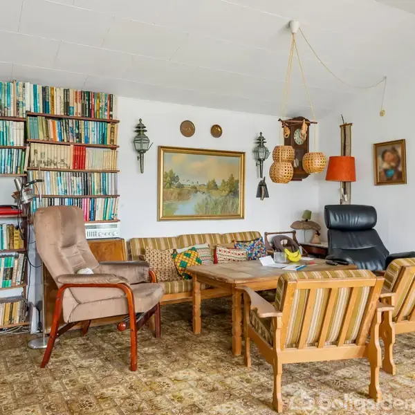 En hyggelig stue med reoler fyldt med bøger langs væggen. Et sofabord er omgivet af blandede møbler, inklusive stole og en sofa. Væggene er dekoreret med malerier og lamper.