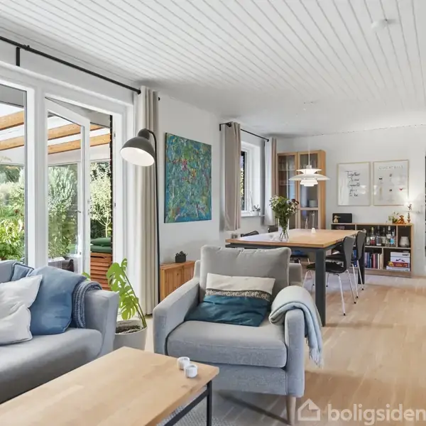 Stue med to sofaer ved et sofabord, vendt mod store vinduer, udsigt til terrasse. Lys træindretning og kunst på væggen. Spisebord i baggrunden.