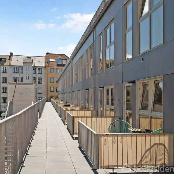 Gangbro med altaner til højre, set i en urban boligkontekst med bygningsfacader i baggrunden under en blå himmel.