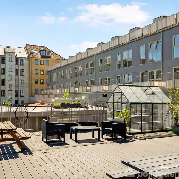 Terrasse med bordbænkesæt og loungemøbler. En lille drivhus står ved siden af plantekrukker. Omkranset af lejlighedsbygninger med blå himmel og spredte skyer i baggrunden.