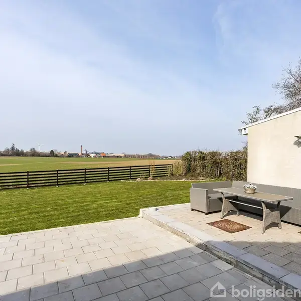 Et fladt bord-sæt står på en terrasse med fliser. Terrassen grænser op til en velplejet græsplæne, der strækker sig mod et åbent marklandskab med en lav træhegn i baggrunden.