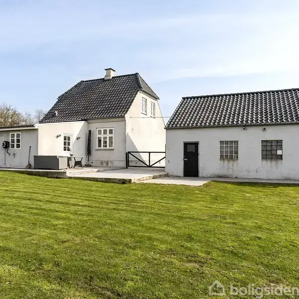Et hvidt hus med sorte tagsten står på en velplejet græsplæne. Det har flere sektioner og en terrasse. Omgivet af træer og en blå himmel i baggrunden.