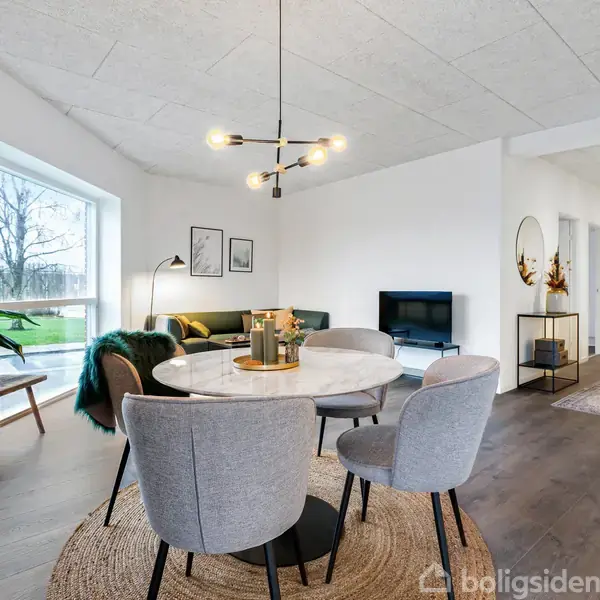 Et moderne stueområde med runde spisebord omgivet af grå stole, sofagruppe i baggrunden, plantelamper, store vinduer til venstre og lysindfald. Decorativt tæppe dækker trægulvet.