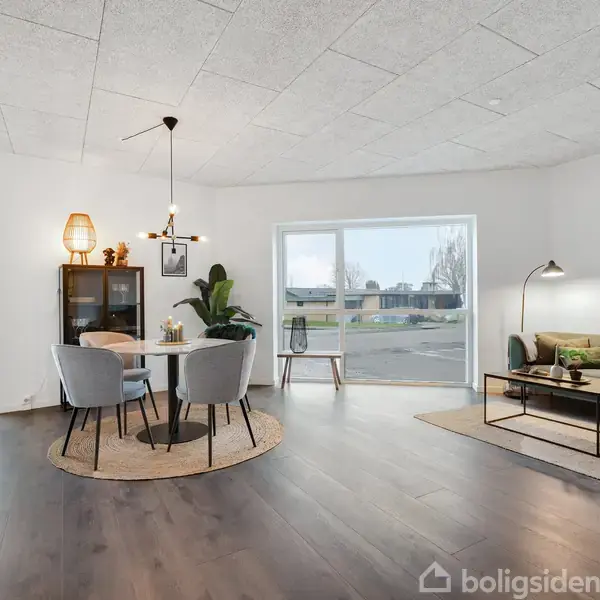 Stue med rundt spisebord, fire stole og en grøn sofa. Store vinduer giver udsigt til et udendørsområde. En reol med planter og lamper skaber en hyggelig atmosfære.