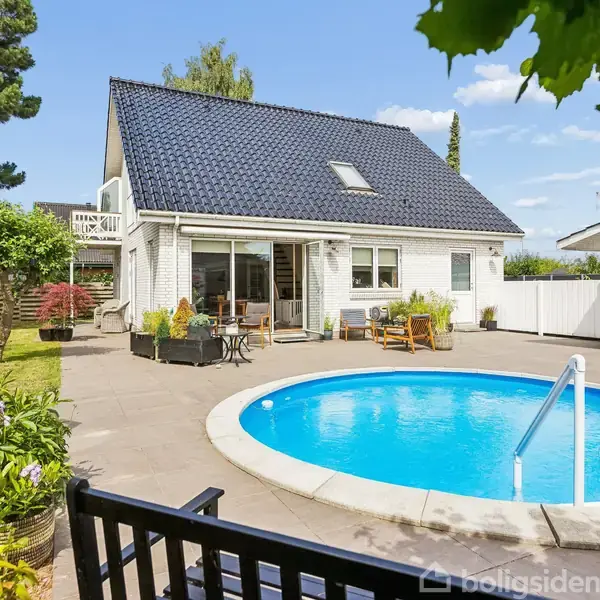 Hus med sort tag ligger ved en terrasse. Rund pool med stige i forgrunden. Omkring poolen er der havemøbler og blomster i krukker.