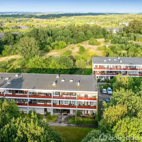Flere boligblokke står stille omgivet af frodig natur og åbne græsarealer. Skove og marker strækker sig i baggrunden under en blå himmel.