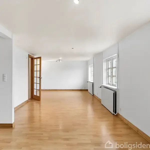 Et lyst, rummeligt værelse med trægulv og hvidmalede vægge. Flere vinduer langs højre væg giver naturligt lys. I baggrunden ses en delvist åben dør til køkkenet.