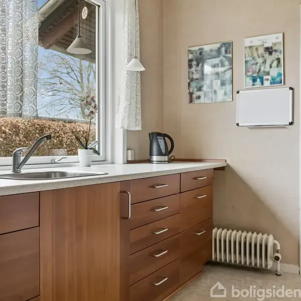 Brunt køkken med bordplade i sten, vask, elkedel og skabslåger. Vindue med blondegardiner lader lys ind og udsigt til træer. Radiator og opslagstavler på væggen. Ingen synlig tekst.