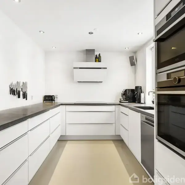 Et moderne køkken med hvide skabe og gråt bordplade. En indbygget ovn og opvaskemaskine ses til højre. Knive hænger på væggen. Rummet er godt oplyst med spotlys.