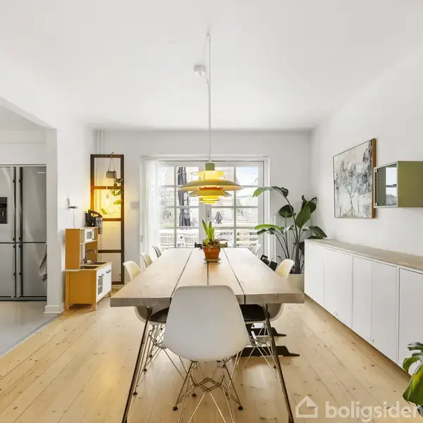 Et lyst spisebord med stole omringes af en moderne spisestue. En hængelampe lyser over bordet. Til venstre ses et åbent køkken, og planter pynter rummet.