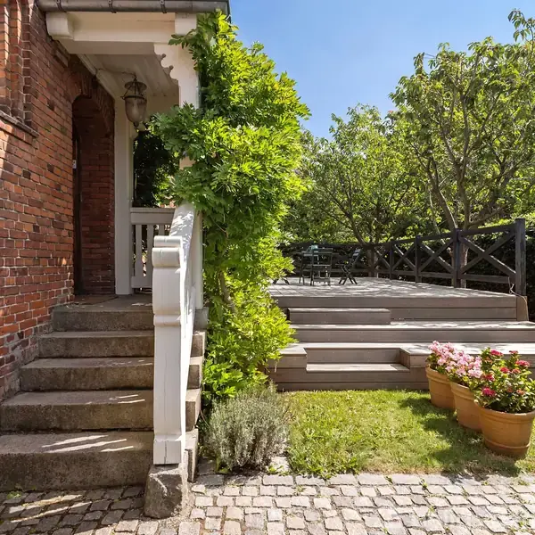 Trappe lavet af mursten fører op til en veranda omgivet af grønne planter. Ved siden af er en træterrasse med havemøbler, omkranset af hække og træer under en klar himmel.