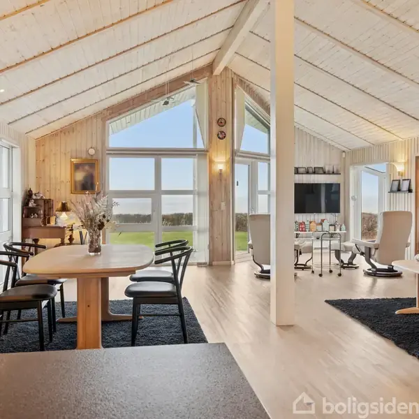 Et lyst og moderne stuerum med træmøbler og sort interiør, omgivet af store vinduer, der giver udsigt til en græsplæne udenfor, og en tv-skærm opstillet i hjørnet.